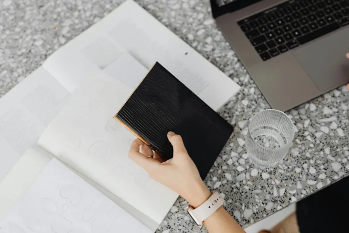 A person holding a black notebook with an artistic design, surrounded by documents and a laptop, indicating a focus on organization and creative work.