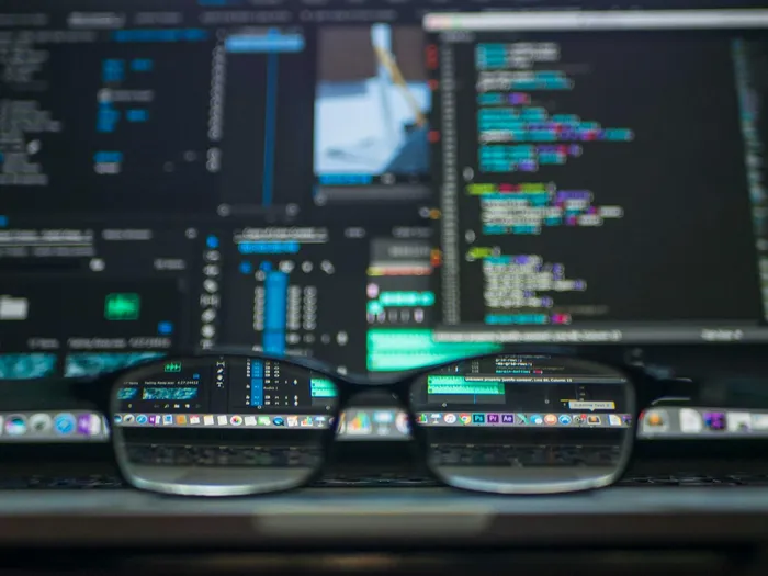 A close-up of eyeglasses in front of a computer screen displaying programming code and software applications.
