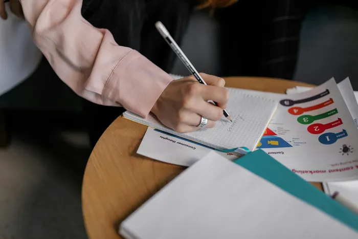 A person writing notes in a notebook while sitting at a table, with documents spread out, indicating a focus on organizing thoughts and ideas.