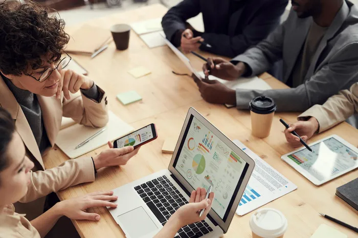 A diverse group of professionals engaged in a collaborative meeting, discussing data displayed on a laptop and presenting insights from various devices.